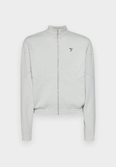 Ανοιχτό γκρι κοντό τζάκετ με φερμουάρ και ψηλό γιακά, τεχνοτραφή ύφασμα, πλαϊνές ραφές και μικρό λογότυπο στην αριστερή περιοχή του στήθους.
