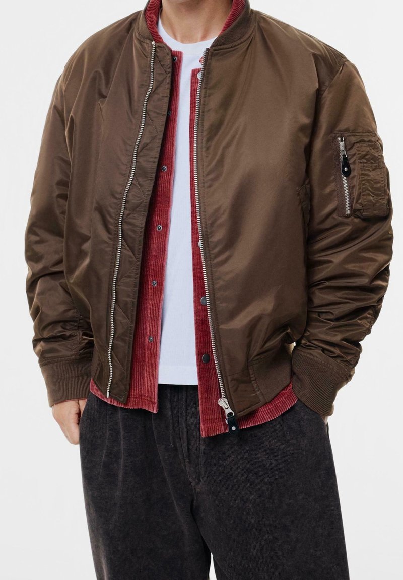 Homme portant une veste bomber marron foncé sur une chemise en velours rouge et un t-shirt blanc, avec un pantalon gris foncé et les mains dans les poches.