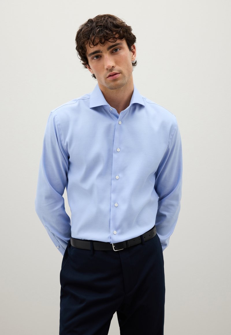 Chemise bleu clair à manches longues avec un col structuré et des boutons sur le devant. Associée à un pantalon foncé et une ceinture noire, présentée sur un fond neutre.