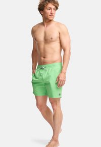 Ενήλικας άντρας με ατημέλητα μαλλιά, φορώντας ανοιχτό πράσινο μαγιό σορτς, με τα χέρια στις τσέπες, όρθιος με γυμνά πόδια μπροστά σε ένα απλό λευκό φόντο.