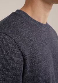 Pull-over en laine marine avec un motif de tricot texturé, un col rond côtelé et un tissu lisse. Vue en gros plan des détails de la manche.