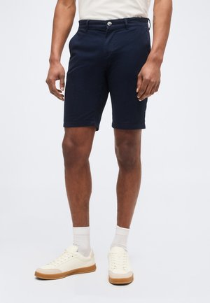 Homme portant un short bleu marine, des chaussettes blanches et des baskets blanches, debout devant un fond clair uni.