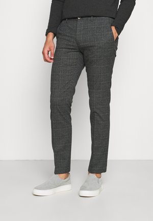 Mann trägt dunkelgraue karierte, schlanke Hose, schwarzen Langarm-Top und hellgraue Slip-On-Sneaker und steht mit einer Hand in der Tasche.