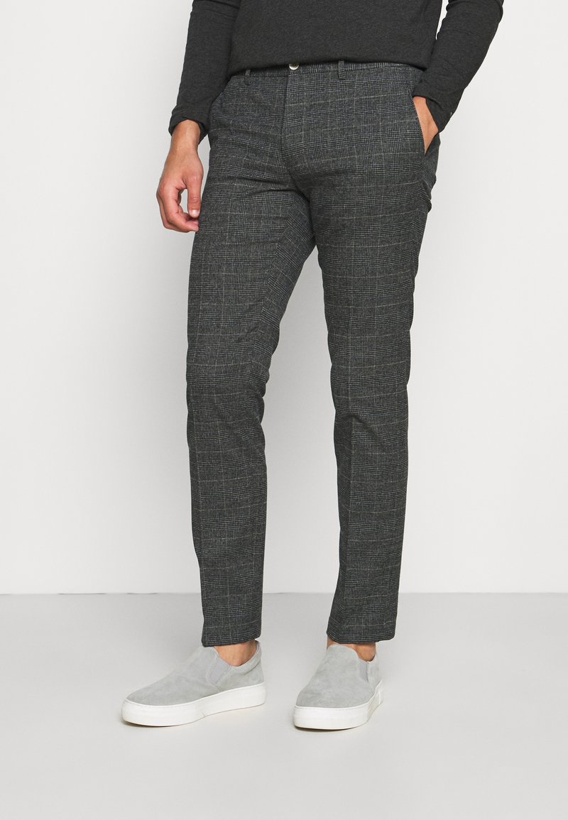 Homme portant un pantalon slim gris foncé à carreaux, un haut noir à manches longues et des baskets gris clair sans lacets, debout avec une main dans la poche.