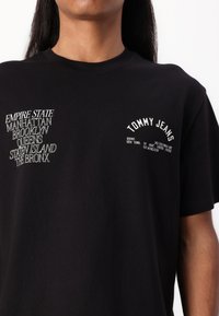 Sort bomulds T-shirt med hvid grafisk tekst. Foran til venstre står der "TOMMY JEANS", og til højre listes NYC-bydelene i varierende stilarter.