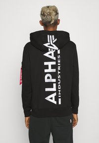 Crna dukserica s kapuljačom s bijelim logotipom "ALPHA INDUSTRIES" na leđima, teksturirana tkanina, podesiva kapuljača i istaknuta crvena oznaka na rukavu.