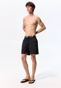 Pantaloni da bagno neri con elastico in vita e logo. Realizzati in tessuto leggero, lunghi fino al ginocchio, con un design semplice.
