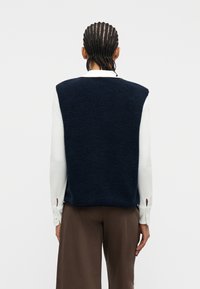 Άτομο με πλεξούδες που φοράει σκούρο μπλε γιλέκο από φλις πάνω από λευκό πουκάμισο με μακριά μανίκια και καφέ παντελόνι με φαρδιά πόδια, κοιτάζοντας προς τα πίσω.