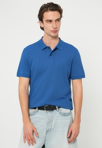 Μπλε βαμβακερό πόλο πουκάμισο με γιακά, κοντά μανίκια και μικρό λογότυπο στο στήθος. Συνδυάζεται με ανοιχτό μπλε τζιν παντελόνι και μαύρη ζώνη.