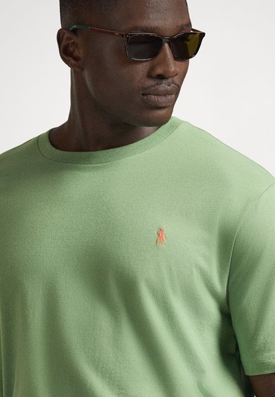 Ανοιχτοπράσινο βαμβακερό T-shirt με ριμπ λαιμόκοψη, διακοσμημένο με κεντημένο πορτοκαλί λογότυπο στο στήθος. Το μοντέλο φοράει σκούρα γυαλιά ηλίου.