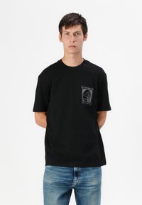 Black cotton t-shirt with a round neck, short sleeves, and a grey graphic pocket print. Paired with light blue denim jeans.