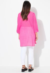 Roze linnen tuniek met driekwartmouwen, zijsplitten en een lichte textuur. Gepaard met witte broeken en zwarte sandaaltjes met bandjes.