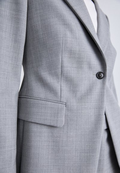 Visão aproximada de uma pessoa vestindo um blazer xadrez cinza claro abotoado uma vez, mostrando o lapel, a aba do bolso e os detalhes da costura lateral.
