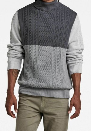 Mężczyzna ubrany w dwukolorowy szary, bezrękawnik z grubym splotem na tle jasnoszarej koszuli z długim rękawem i beżowe spodnie, stojący z rozluźnionymi ramionami.