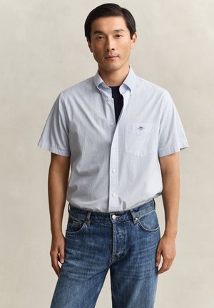 Mann trägt hellblaues kurzärmeliges Hemd über schwarzem T-Shirt und blaue Jeans, steht vor einem einfarbigen beigen Hintergrund.