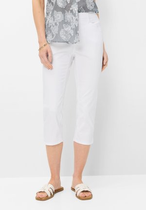 Frau mit weißen, gekürzten Hosen, gemustertem transparentem Oberteil, goldenem Armband und weißen Slip-On-Sandalen vor schlichtem Hintergrund stehend.