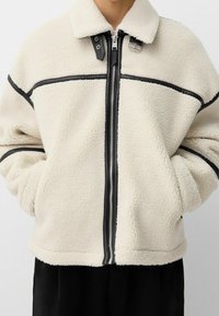 Krämfärgad fleecejacka med svarta läderdetaljer, dragkedja och krage. Har en avslappnad passform och en texturerad yta.