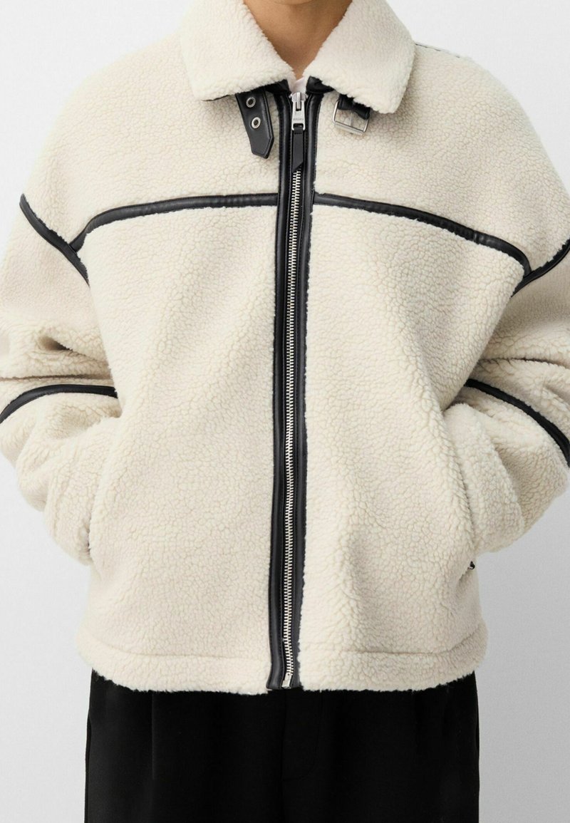 Krämfärgad fleecejacka med svarta läderdetaljer, dragkedja och krage. Har en avslappnad passform och en texturerad yta.
