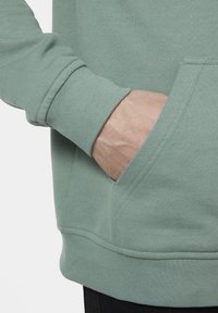 Sudadera de color verde claro con un bolsillo tipo canguro y puños acanalados. Textura suave, con costuras dobles en el dobladillo y los bordes del bolsillo.