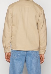 Beige bomullsjacka med långa ärmar, synliga sömmar, knäppning vid ärmsluten och rak fåll, ihop med ljusblå jeans.