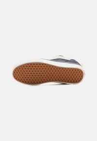 Suela de zapatilla con una parte inferior de goma texturizada en marrón con un patrón de diamante; paredes laterales blancas y materiales superiores en azul oscuro.