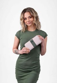 Světle růžová peněženka s vzorovaným černým a zeleným pruhem, držena osobou v zelených šatech; hladký povrch a obdélníkový tvar.