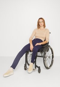 Light beige ribbed long-sleeve top, navy blue tailored pants with buttons, beige slip-on shoes, and a wheelchair on a plain background.