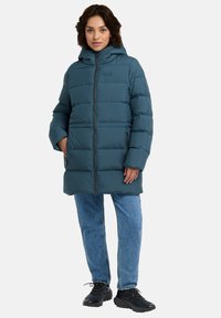 Giacca a vento teal con cappuccio, caratterizzata da cuciture orizzontali, due cerniere laterali e una cerniera frontale. Abbinata a jeans azzurri chiari e scarpe nere.