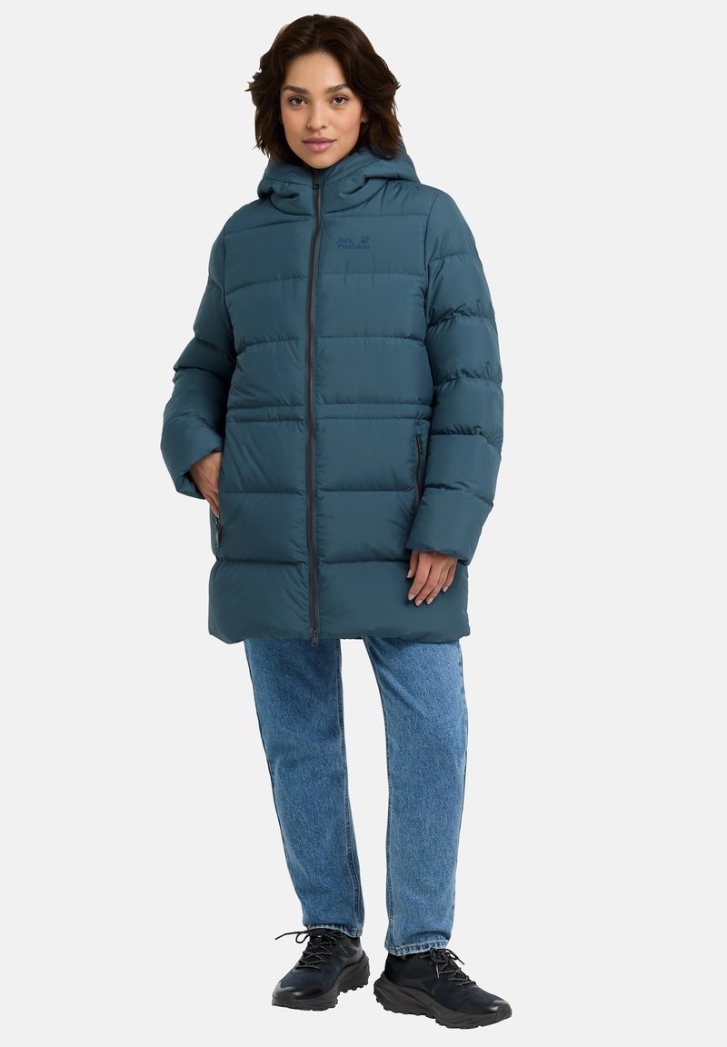 Giacca a vento teal con cappuccio, caratterizzata da cuciture orizzontali, due cerniere laterali e una cerniera frontale. Abbinata a jeans azzurri chiari e scarpe nere.