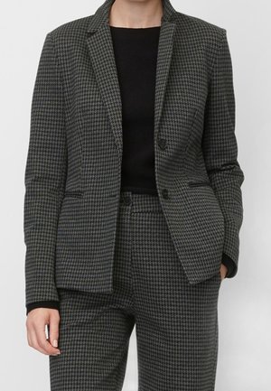 Femme portant un blazer pied-de-poule gris charbon et un pantalon assorti avec un top noir en dessous, mains détendues sur les côtés.