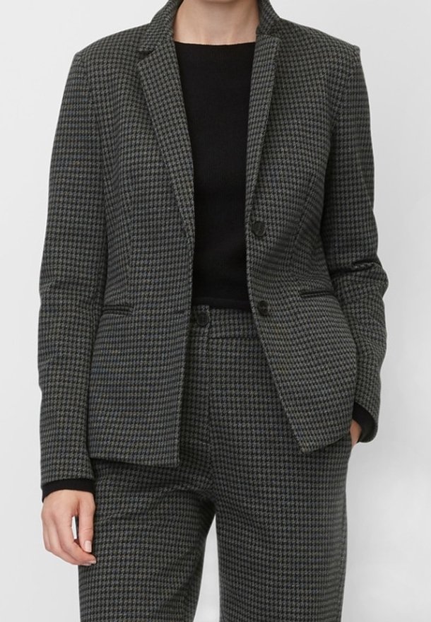 Femme portant un blazer pied-de-poule gris charbon et un pantalon assorti avec un top noir en dessous, mains détendues sur les côtés.