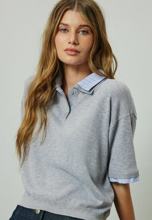 Ung kvinde med langt lysebrunt hår iført en lysegrå kortærmet sweater over en blåstribet skjorte med krave.