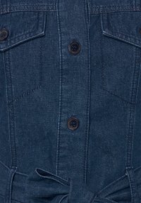 Mörkblå denimplagg med knäppning, två bröstfickor och ett knytbart tygbälte i midjan.