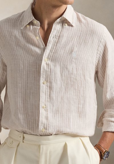 Homme portant une chemise beige clair rayée à boutons avec manches retroussées et un pantalon plissé crème, main dans la poche, portant une montre marron.