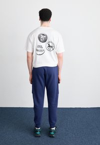 Biała koszulka z trzema czarnymi okrągłymi grafikami na plecach, zestawiona z granatowymi spodniami dresowymi i obuwiem z akcentami w kolorze turkusowym.