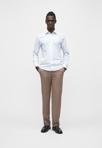 Homme debout, mains dans les poches, portant une chemise blanche à boutons avec un motif floral bleu, un pantalon beige et des chaussures marron foncé.