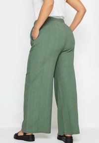 Zelené široké kalhoty s texturovanou tkaninou, obsahující boční kapsy a vysoký elastický pas, ve spojení s černými platformovými sandály.