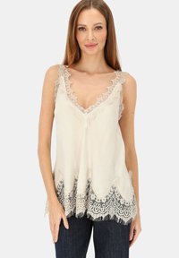 Kremowy, bezrękawnik z głębokim dekoltem w kształcie litery V, ozdobiony misterną koronką, falistym dołem i frędzlami u podstawy, uzupełniony przezroczystą teksturą.