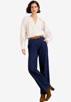Blusa bianca ricamata con scollatura a V e maniche lunghe, abbinata a pantaloni a gamba larga blu scuro e stivaletti con tacco marroni.