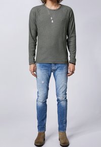 Chemise texturée vert olive à manches longues, associée à un jean bleu clair déchiré et des bottines en daim marron. Collier superposé simple visible.