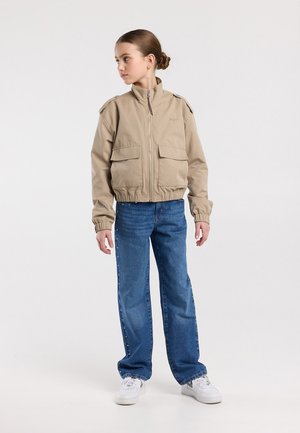 Mädchen mit Dutt, das eine beige Jacke mit Reißverschluss, blaue Jeans mit seitlichen Nieten und weiße Turnschuhe trägt und nach links schaut.