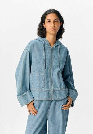 Hellblaues Jeansjacke mit Kapuze, Frontreißverschluss und zwei großen Taschen; hat weite Ärmel und sichtbare Nähte.