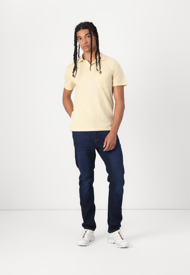 Camisa polo amarilla claro con un bolsillo, combinada con jeans azul oscuro y zapatillas blancas. El modelo está de pie frente a un fondo blanco liso.