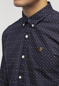 Granatowa koszula zapinana na guziki z wzorem w kropki w kolorze białym, niebieskim i rdzy, z małym haftowanym logo na piersi.