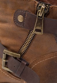 Botín de cuero marrón hasta el tobillo con un zipper lateral, una correa decorativa y acentos de hardware de latón. Superficie texturizada y costuras visibles.