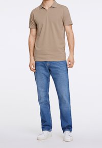 Muž oblečený v světle hnědém polo tričku s krátkým rukávem, modrých džínách a bílých teniskách, stojící proti čistému bílému pozadí.