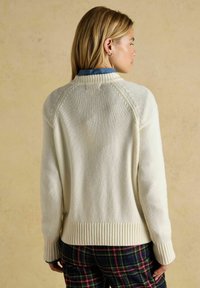 Krēmkrāsas adīta džemperis ar ribotu apakšmalu un aprocēm, ar raglāna piedurknēm un strukturētu adījuma rakstu. Nēsāts virs zila apkakles krekla.
