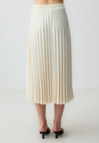 Gelaagde midi-jurk in crèmekleurige stof met een elastische tailleband. Heeft een zachte textuur en vloeiende draping, rustend boven zwarte hakken.