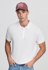 Biała koszulka polo z klasycznym kołnierzykiem, dwoma guzikami oraz podwiniętymi rękawami, w połączeniu z jasnoniebieskimi dżinsami i burgundową czapką baseballową.