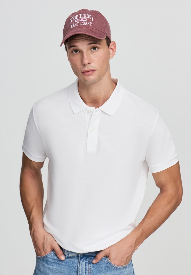 Biała koszulka polo z klasycznym kołnierzykiem, dwoma guzikami oraz podwiniętymi rękawami, w połączeniu z jasnoniebieskimi dżinsami i burgundową czapką baseballową.